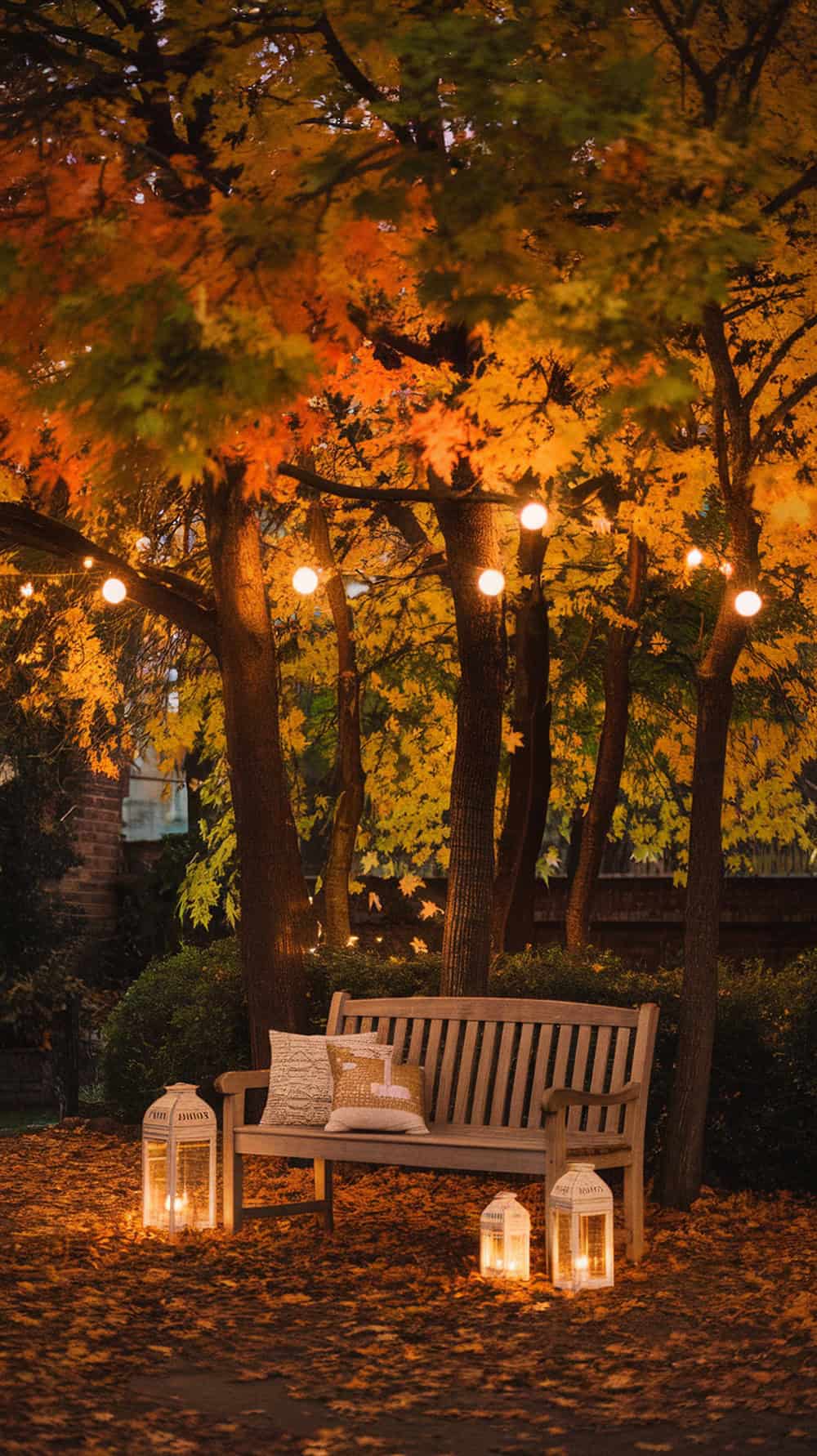 A wooden bench with pillows is surrounded by autumn leaves and illuminated by string lights hanging from trees above. Lanterns with candles on the ground add a warm, cozy ambiance to the scene.