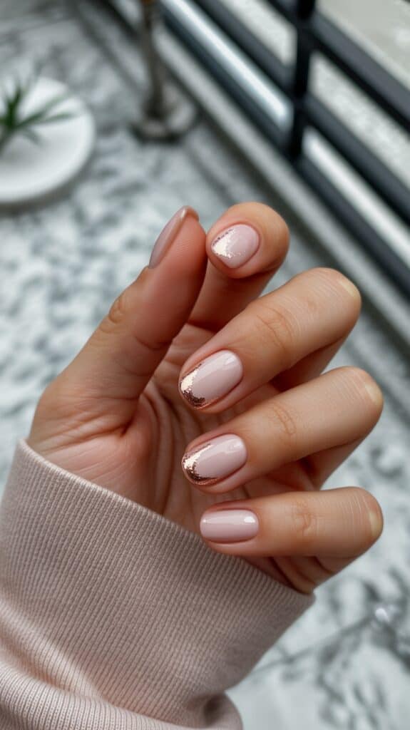 A hand with a manicure featuring light pink nails polished with a metallic, glittery rose gold tip design. The hand is held in a relaxed manner, and the person is wearing a pink long-sleeved shirt. The background includes a blurred marble surface and a window with bars.