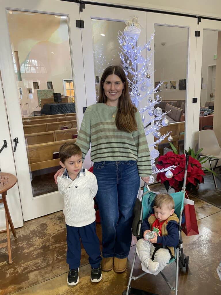 A woman stands smiling with two children, one beside her and one in a stroller, in front of a white decorated tree.