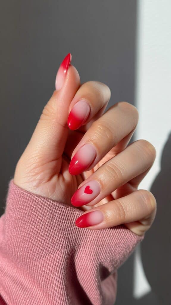 A hand with a manicure featuring a red gradient effect on almond-shaped nails, with a small red heart design on one nail. The person is wearing a pink sweater with their sleeve partially visible.